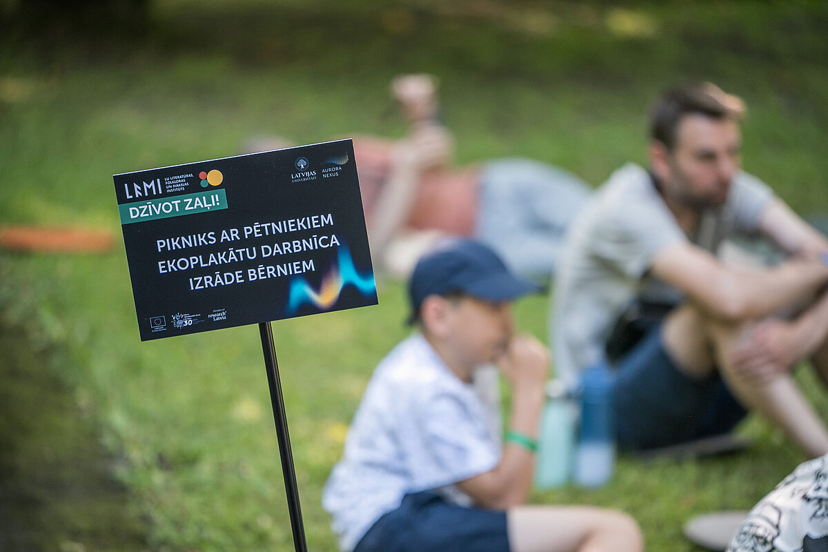 Foto: Toms Grīnbergs, LU Melna informatīva zīme zālienā aicina uz pasākumu “Dzīvot zaļi!”, norādot aktivitātes – pikniku ar pētniekiem, ekoplakātu darbnīcu un izrādi bērniem. Fonā parkā uz zāles atpūšas pasākuma apmeklētāji, tostarp bērni un pieaugušie, bet attēls fokusēts uz zīmi.