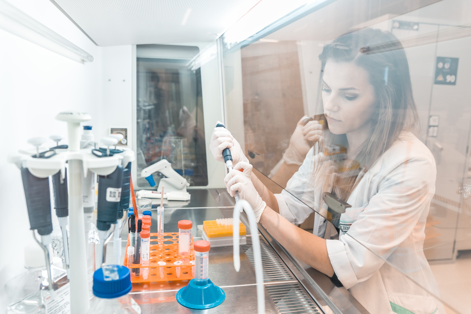 Laboratorijas telpa ar aizsargstiklu un darba virsmu; persona laboratorijas halātā un cimdos ar pipeti strādā ar mēģenēm stendā, uz galda redzams laboratorijas aprīkojums, pipetes un trauki ar paraugiem.