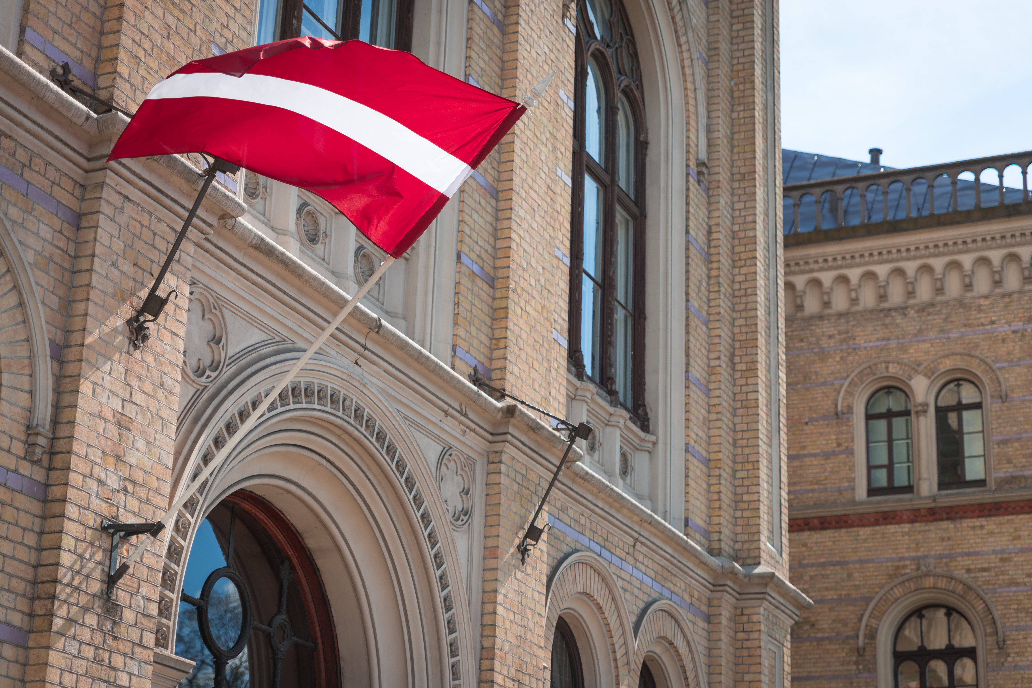 Latvijas karogs plīvo pie vēsturiskas ķieģeļu ēkas fasādes ar arku durvīm un dekoratīviem akmens ornamentiem - Latvijas Universitātes galvenā ēka Rīgā